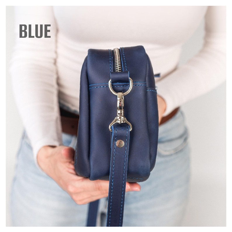 May include: A blue leather crossbody bag with a silver zipper and strap hardware. The bag has a long, adjustable strap and the word "BLUE" is printed in the upper left corner. The bag is held by a person.
