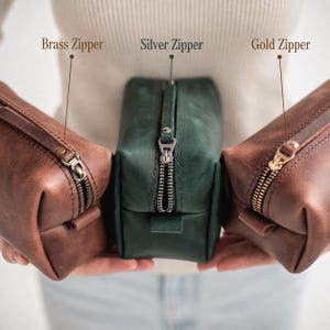 May include: Three leather toiletry bags in brown, green, and brown, each with a different zipper color: brass, silver, and gold. The bags are rectangular with a zipper closure and a small leather loop on one side. The text above the bags reads "Brass Zipper", "Silver Zipper", and "Gold Zipper".