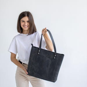 May include: A black leather tote bag featuring a large rectangular front pocket, two long shoulder straps attached with silver-toned metal rivets, and a top zipper closure. The bag has a matte finish with visible natural texture and grain.