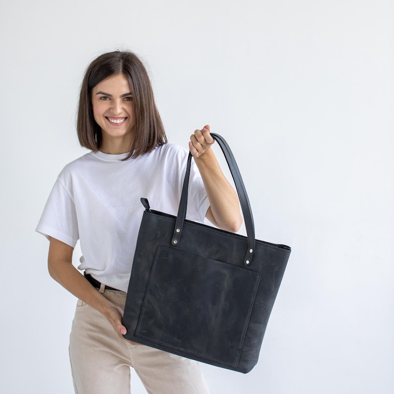 May include: A black leather tote bag featuring a large rectangular front pocket, two long shoulder straps attached with silver-toned metal rivets, and a top zipper closure. The bag has a matte finish with visible natural texture and grain.