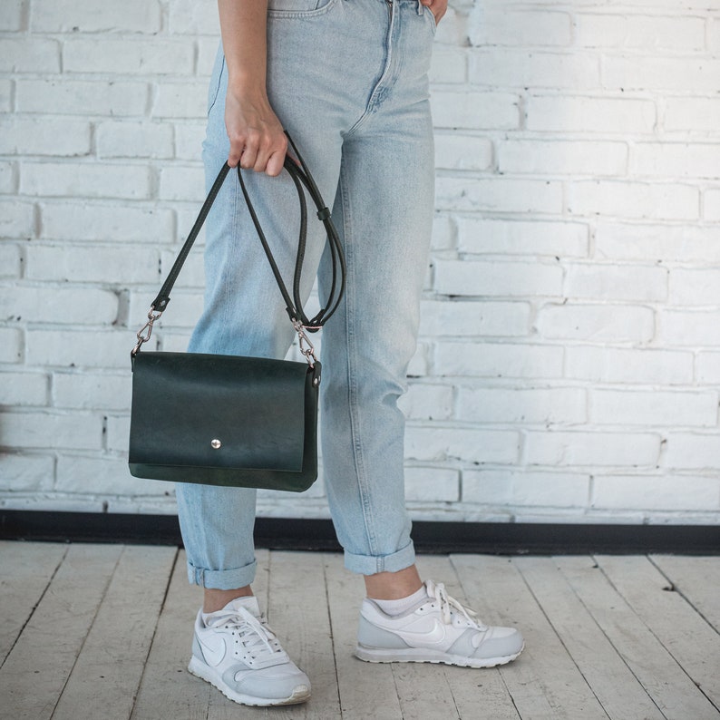 Pode incluir: Uma bolsa transversal de couro verde escuro com fecho de aba e uma al&ccedil;a longa e ajust&aacute;vel. A bolsa &eacute; segurada por uma pessoa que usa jeans azul claro e t&ecirc;nis brancos. O fundo &eacute; uma parede de tijolos brancos.