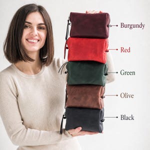 May include: A stack of five leather pouches in various colors: burgundy, red, green, olive, and black. Each pouch has a zipper closure and a wrist strap. The pouches are held up by a person.