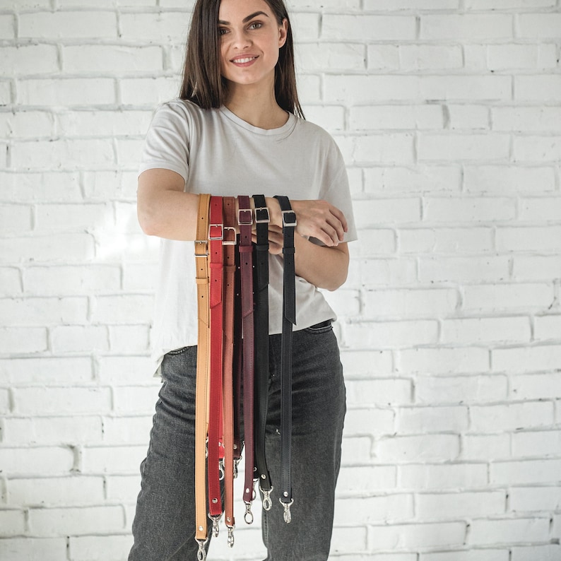 Pode incluir: Uma sele&ccedil;&atilde;o de al&ccedil;as de bolsa de couro em v&aacute;rias cores, incluindo bege, vermelho, bord&ocirc; e preto. Cada al&ccedil;a apresenta ferragens prateadas, incluindo fivelas e fechos. As al&ccedil;as s&atilde;o exibidas contra uma parede de tijolos brancos.