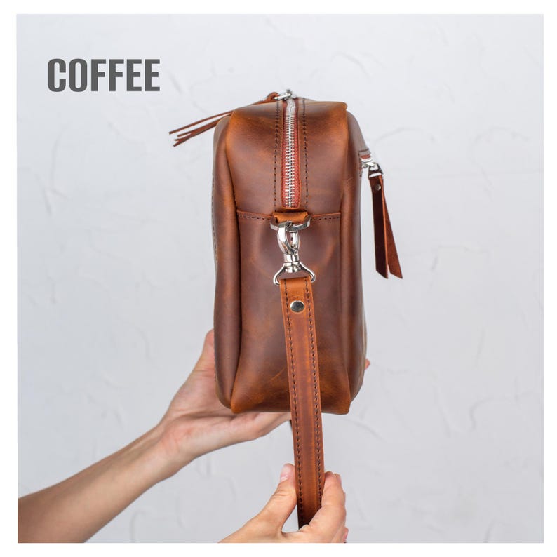 Pode incluir: Uma bolsa transversal de couro marrom com z&iacute;per e al&ccedil;a prateados. A bolsa tem formato retangular e uma pequena al&ccedil;a. A palavra "COFFEE" est&aacute; impressa em cinza no topo.