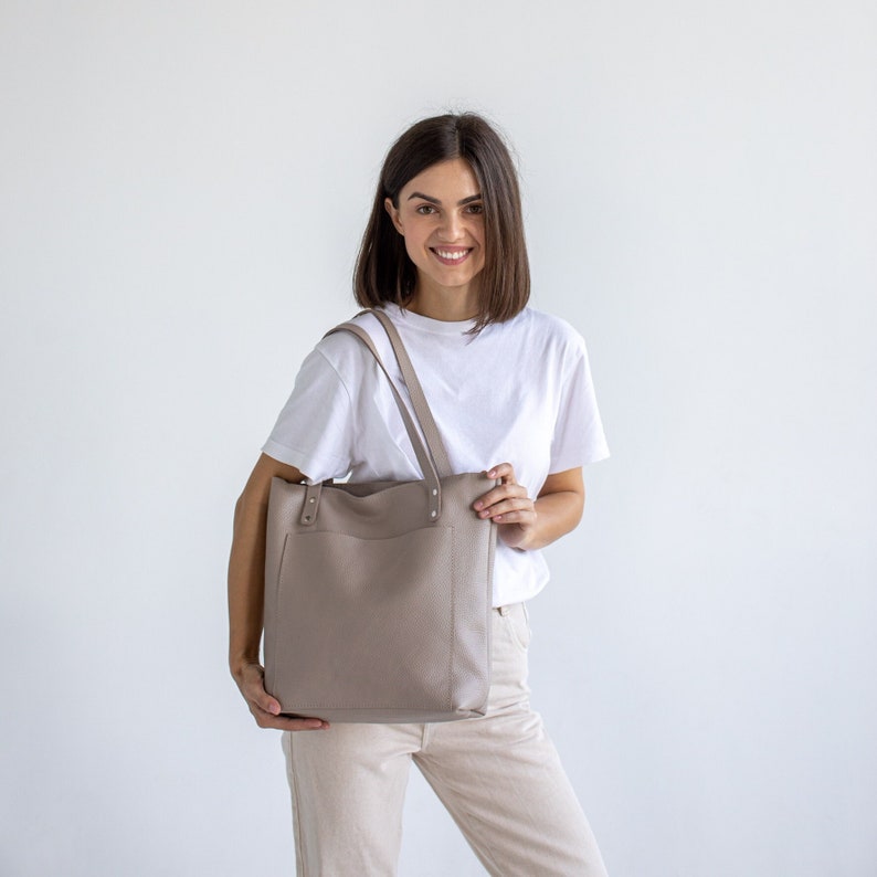 May include: A woman is holding a light brown leather tote bag with a white t-shirt and beige pants.