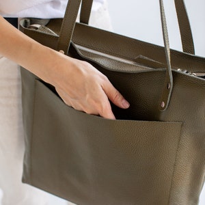 May include: A large, olive green leather tote bag with a zipper closure and a large front pocket. The bag has a textured, pebbled leather finish.