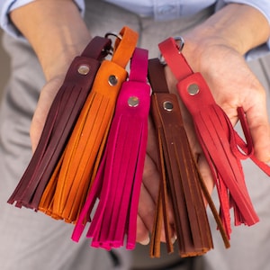 Puede incluir: Una colección de borlas de cuero de colores: burdeos, naranja, fucsia, marrón y rojo. Cada borla tiene un remache plateado y un lazo para sujetar. Las borlas están sostenidas en las manos de una persona.
