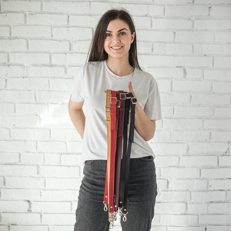 Pu&ograve; includere: Una donna tiene in mano una pila di cinturini per fotocamera in pelle colorati con fibbie argentate. I cinturini sono in tonalit&agrave; di marrone, rosso e nero.