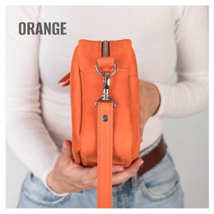Pode incluir: Bolsa transversal de couro laranja com z&iacute;per e ferragens prateadas. A bolsa tem uma al&ccedil;a longa e remov&iacute;vel. A palavra "ORANGE" est&aacute; impressa em branco na parte superior da imagem. A bolsa &eacute; segurada por uma pessoa.