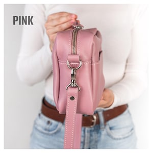 Pode incluir: Uma bolsa transversal de couro rosa com z&iacute;per e ferragens de al&ccedil;a prateadas. A bolsa tem formato retangular e uma al&ccedil;a remov&iacute;vel. A palavra "PINK" est&aacute; impressa no canto superior esquerdo.