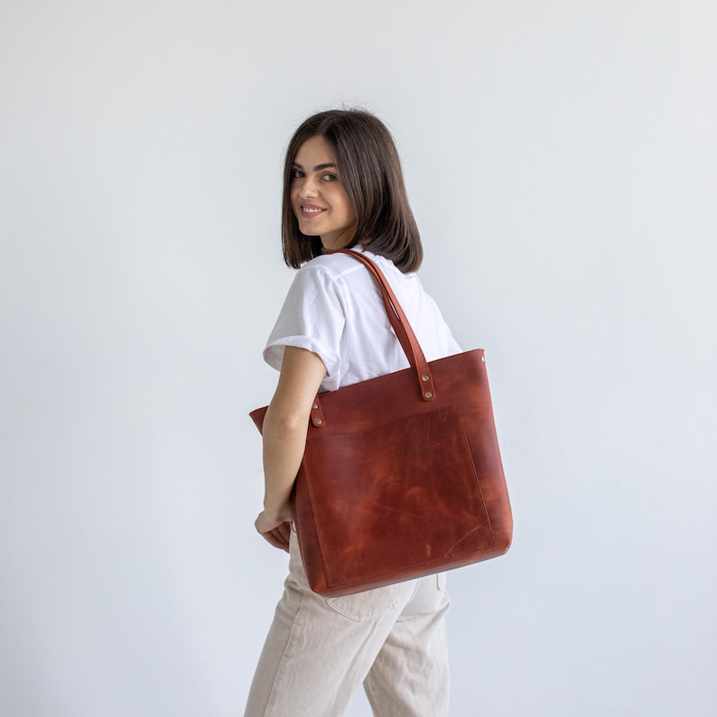 May include: A large, rectangular, brown leather tote bag with long straps. The bag has a pocket on the front and is carried over the shoulder. The leather has a natural, textured appearance.