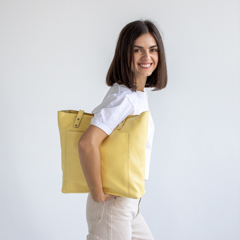 May include: A woman is holding a yellow leather tote bag with a front pocket. The bag has a top zipper closure and two handles.