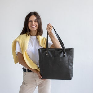 May include: A woman is holding a black leather tote bag with a front pocket. The bag has a top handle and is made of a textured material.