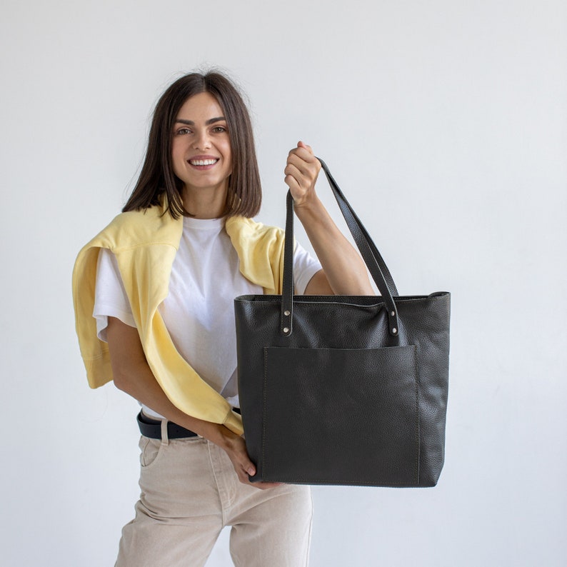 May include: A woman is holding a black leather tote bag with a front pocket. The bag has a top handle and is made of a textured material.