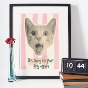 Könnte beinhalten: Ein gerahmter Kunstdruck mit einer Katzenkopf-Illustration im Rasterverfahren, vor einem Hintergrund aus rosa und cremefarbenen Streifen. Der Druck enthält den Text "It's okay to fail. Try again". Eine rote Blume in einer blauen Vase und eine schwarze Klappuhr sind ebenfalls zu sehen.