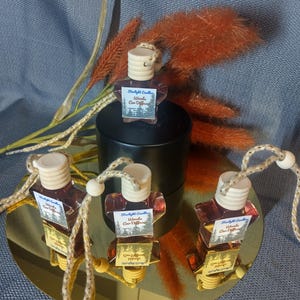 Puede incluir: Cuatro ambientadores para coche con tapones de madera y cordones colgantes se exhiben sobre una superficie dorada. Cada botella contiene un líquido marrón rojizo y una etiqueta que dice "Woods Car Diffuser". El fondo incluye un cilindro negro y plantas decorativas naranjas.