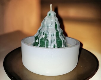 Puede incluir: Una vela cónica de color verde oscuro con gotas de cera blanca que parecen nieve, colocada dentro de un recipiente redondo, blanco y moteado. La vela tiene una mecha central en la parte superior, creando un aspecto texturizado y en capas.