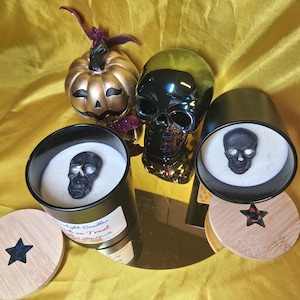 May include: A Halloween-themed arrangement featuring a gold pumpkin, a black skull, and two black candles with skull accents. The candles have wooden lids with star inlays. The background is a yellow fabric.