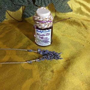 May include: A small glass bottle labeled "Starlight Candles Lavender Car Diffuser" with a decorative rope and cork top. The bottle sits on a yellow surface with dried lavender sprigs. The label indicates the fragrance is lavender.