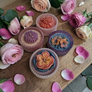 May include: A collection of floral-shaped candles in various colors, including pink, orange, and purple, arranged on a wooden surface. The candles are surrounded by fresh roses and scattered petals, creating a decorative and aromatic display.