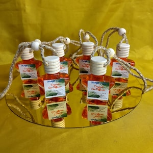 May include: A collection of orange car air fresheners with white wooden caps and cords, displayed on a mirrored surface. Each bottle is labelled "Tropical Car Diffuser". The background is a plain yellow.