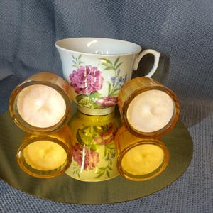 Könnte beinhalten: Eine weiße Teetasse mit einem Blumendesign mit rosa Blüten und grünen Blättern. Vier kleine, bernsteinfarbene Glas-Kerzenhalter mit brennenden Kerzen sind um die Teetasse auf einer reflektierenden Oberfläche angeordnet. Die Kerzen haben einen weichen, warmen Schein.