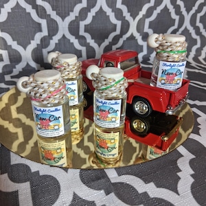 May include: A collection of small glass bottles with the text "New Car" on the label, arranged on a gold-colored tray. The bottles are topped with decorative rope and a small red toy truck is in the background.