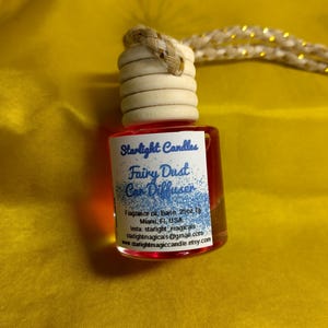 Puede incluir: Un pequeño difusor de coche de vidrio transparente lleno de líquido rojo. La etiqueta dice "Starlight Candles Fairy Dust Car Diffuser". La botella tiene una tapa de madera y una cuerda trenzada para colgar. El fondo es una tela amarilla brillante.