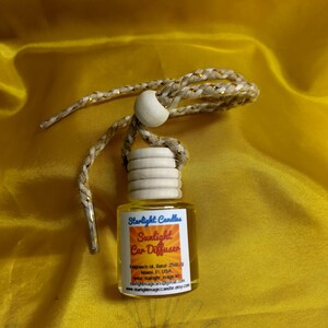 Può includere: Un piccolo flacone di profumo per auto in vetro trasparente con un tappo di legno e un cordino intrecciato. L'etichetta recita "Sunlight Car Diffuser" di "Starlight Candles". Il cordino è beige con dettagli dorati.