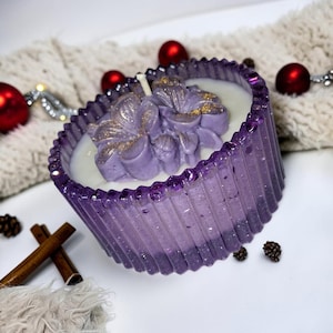 May include: A round, ribbed purple candle container made of translucent resin with embedded glitter. The candle features a white wax base topped with sculpted purple floral wax decorations accented with gold shimmer and a single centered wick.
