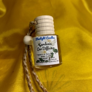 May include: A small glass car diffuser with a wooden cap and a beige string. The label reads "Starlight Candles Gardenia Car Diffuser". The background is a bright yellow fabric.