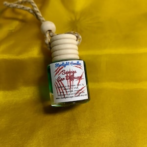 May include: A small, clear glass car air freshener bottle with green liquid and a wooden cap. The label reads "Savage Car Diffuser" with the Starlight Candles logo. It hangs from a beige string.