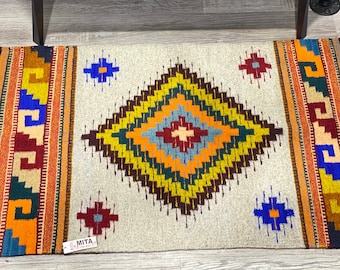 May include: A colorful, handwoven rug with a geometric diamond pattern in shades of orange, yellow, green, blue, and red. The rug has a white background and brown fringe on the edges. The rug is labeled "MITA" in the bottom right corner.