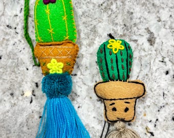 May include: Two felt cactus ornaments with tassels. The cactus on the left is green with a purple flower and a blue tassel. The cactus on the right is green with a yellow flower and a beige tassel.