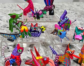 Pode incluir: Uma coleção de alebrijes de madeira pintados com cores vibrantes, incluindo um gato, um cão, um pássaro e outras criaturas fantásticas. Cada figura apresenta padrões intrincados e cores brilhantes como vermelho, verde, azul e roxo, mostrando a arte popular mexicana tradicional.