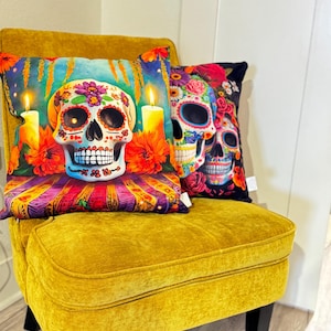 May include: Two decorative throw pillows with a Day of the Dead theme. The pillows feature colorful skull designs with floral accents. The pillows are on a mustard yellow velvet chair.
