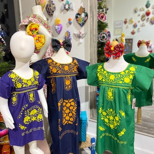 May include: Three embroidered girls' dresses on mannequins. The dresses are in shades of blue, green, and purple with yellow floral embroidery. The dresses are displayed in a shop with a mirror in the background.