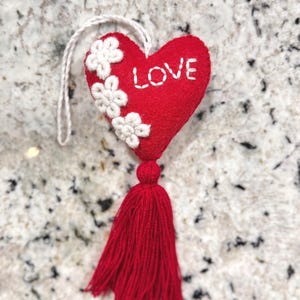 May include: A red heart-shaped ornament with the word "LOVE" embroidered in white. White floral accents adorn the left side. A red tassel hangs below, and a white string is attached for hanging. This decorative item is perfect for Valentine's Day or other special occasions.