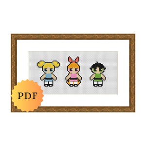 May include: A framed cross-stitch featuring three cartoon characters in a row. The characters are in the style of the Powerpuff Girls, with blonde, orange, and black hair. The characters are wearing dresses in blue, pink, and green. A yellow PDF sticker is in the lower left corner.