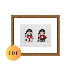May include: Cross-stitch artwork featuring two hockey players in red and black uniforms, each with the number 7. One player holds a hockey stick and the other a hockey stick and puck. The artwork is framed in a gold-coloured wooden frame.