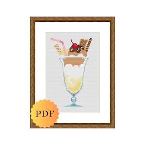 Puede incluir: Un patrón de punto de cruz de un vaso de helado con crema batida, salsa de chocolate, una cereza y una pajita. El patrón está enmarcado en un marco de madera marrón.