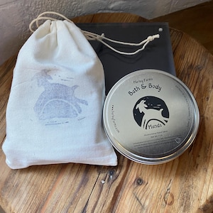 May include: A round silver metal tin of Harley Farms Bath & Body Hands moisturizing hand salve with a net weight of 7 oz, accompanied by a white drawstring fabric pouch featuring a grey goat graphic print on the front.