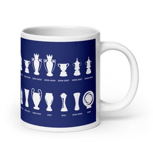 Pode incluir: Caneca de cerâmica branca com fundo azul com silhuetas brancas de troféus e os anos em que foram ganhos. A caneca tem uma alça branca e um acabamento brilhante. Os troféus estão dispostos em fileiras.