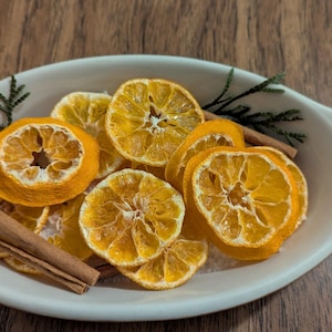 Puede incluir: Un plato ovalado blanco lleno de rodajas de naranja secas, palitos de canela y ramitas verdes. Las rodajas de naranja son de color naranja vibrante y los palitos de canela son de color marrón cálido. El plato está sobre una superficie de madera.
