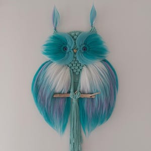 Peut inclure: Une décoration murale en macramé en forme de hibou. Le hibou présente des accents turquoise, blancs et violets. Le corps du hibou est fait de macramé tissé, avec des ailes duveteuses turquoise et un perchoir en bois. Le hibou a deux touffes plumeuses sur la tête.