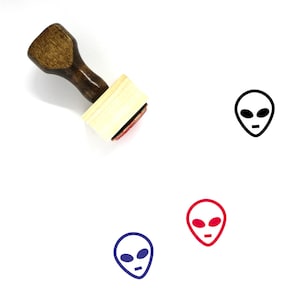 May include: A wooden rubber stamp with a black alien face design. The stamp is on a white background. There are three alien faces in different colors: black, red, and blue.