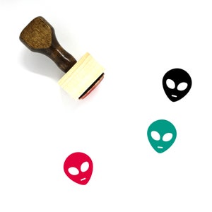 May include: A wooden rubber stamp with a black, red, and green alien head design. The stamp is on a white background.