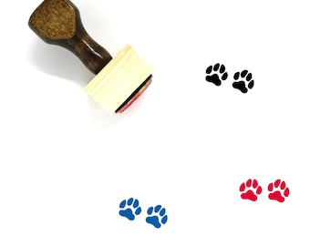 Puede incluir: Un sello de madera con mango y base cuadrada. El sello presenta un diseño de huella de pata. El sello se muestra con tres huellas de pata de diferentes colores: negro, azul y rojo.