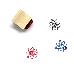 Puede incluir: Un sello de madera con una almohadilla de tinta roja. El sello presenta un dibujo de l&iacute;nea en blanco y negro de un &aacute;tomo con un n&uacute;cleo y electrones en &oacute;rbita. El &aacute;tomo tambi&eacute;n se muestra en rojo y azul.