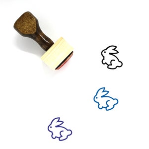 Puede incluir: Sellos de goma de madera con mango marrón y base rectangular. El sello presenta un dibujo lineal simple de un conejo en tres colores diferentes: negro, azul y morado.